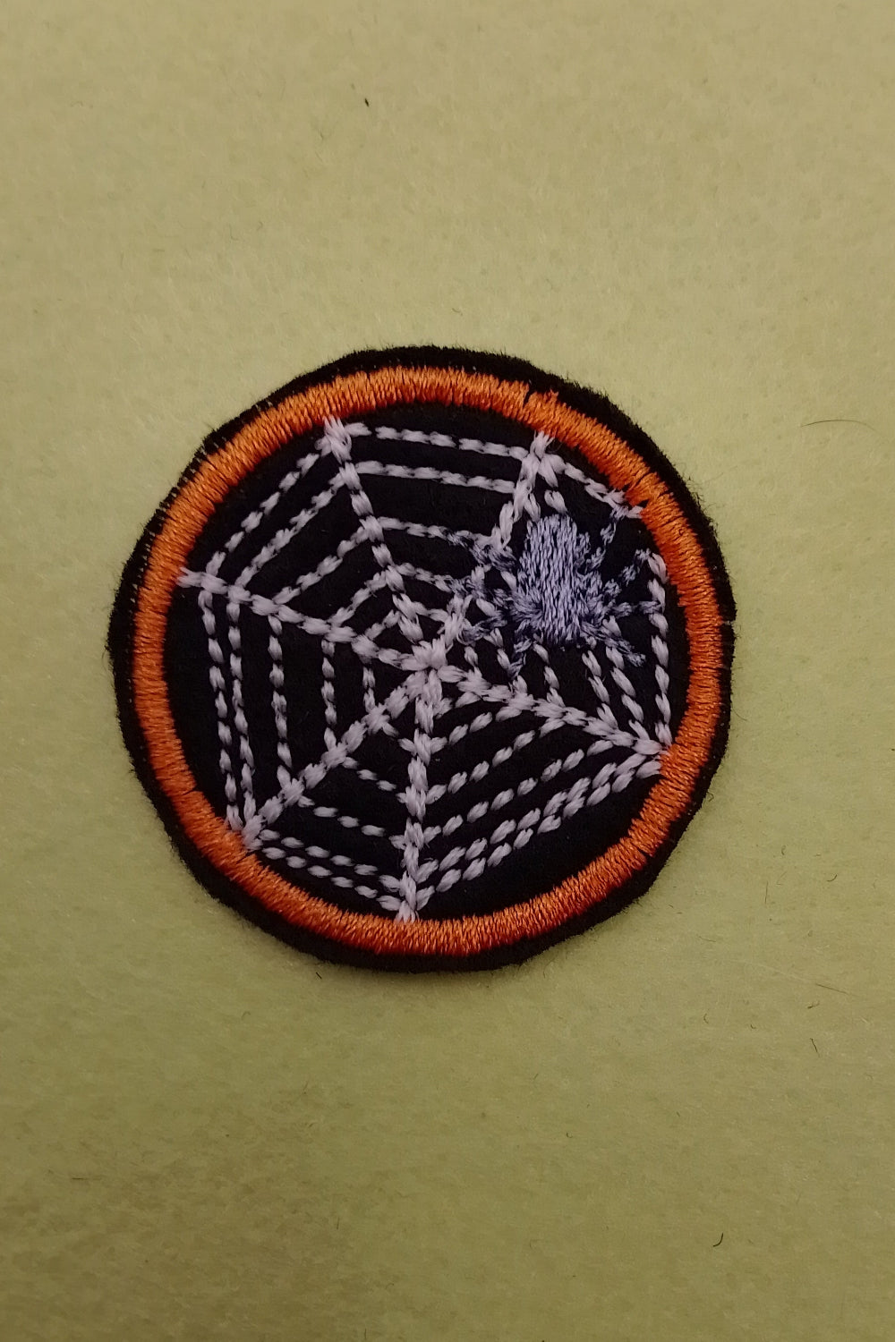 Runde Aufnäher mit orange Außenrand, Spinnennetz das leuchtet im dunkeln und eine kleine graue Spinne